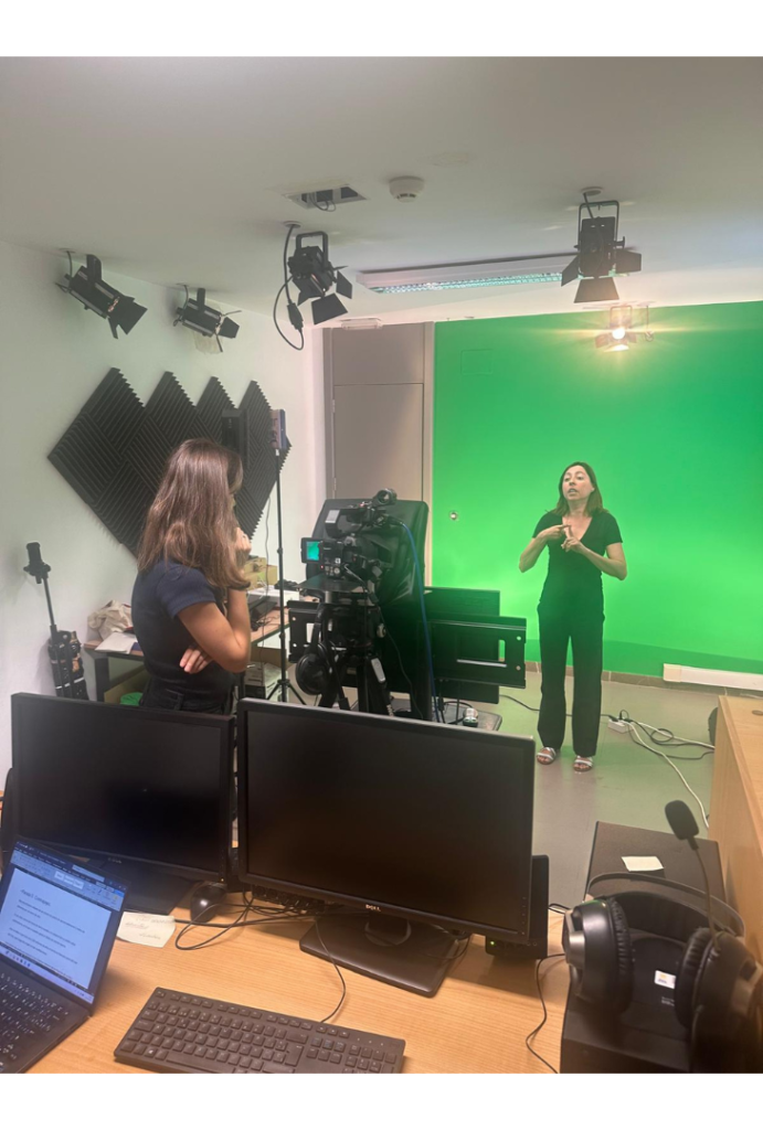 Foto del momento de la grabación en el estudio. En primer plano se ven varias pantallas de ordenador. En el plano medio una persona tras la cámara. En el fondo, la intérprete mientras signa frente a un croma verde de fondo.